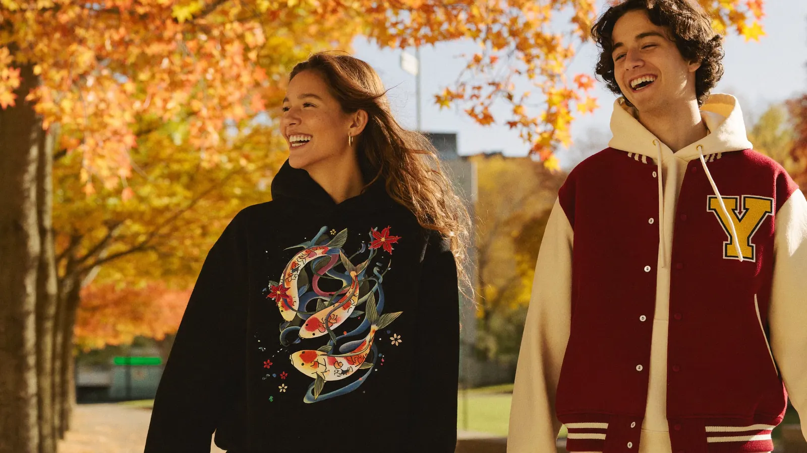 Two friends on an autumn college campus wearing an anime graphic hoodie and a varsity jacket