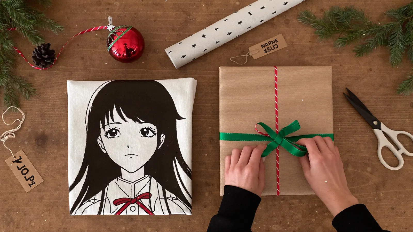 Hands wrapping Christmas gifts with a folded anime graphic tee and an ornament on a wooden table